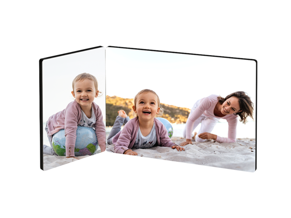 Ein zweiteiliger Fotoaufsteller zeigt eine fröhliche Szene am Strand. Auf der linken Seite sitzt ein kleines Mädchen auf einem bunten Ball und lächelt. Auf der rechten Seite krabbelt ein anderes kleines Mädchen im Sand und schaut lachend zur Kamera. Eine junge Frau kniet daneben im Sand und lächelt ebenfalls. Im Hintergrund ist ein heller Sandstrand und etwas Grün zu sehen.